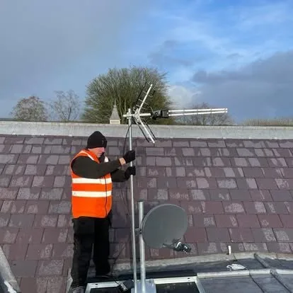 An engineer in hi-vis on a slate roof, aligning an aerial beside a satellite dish.